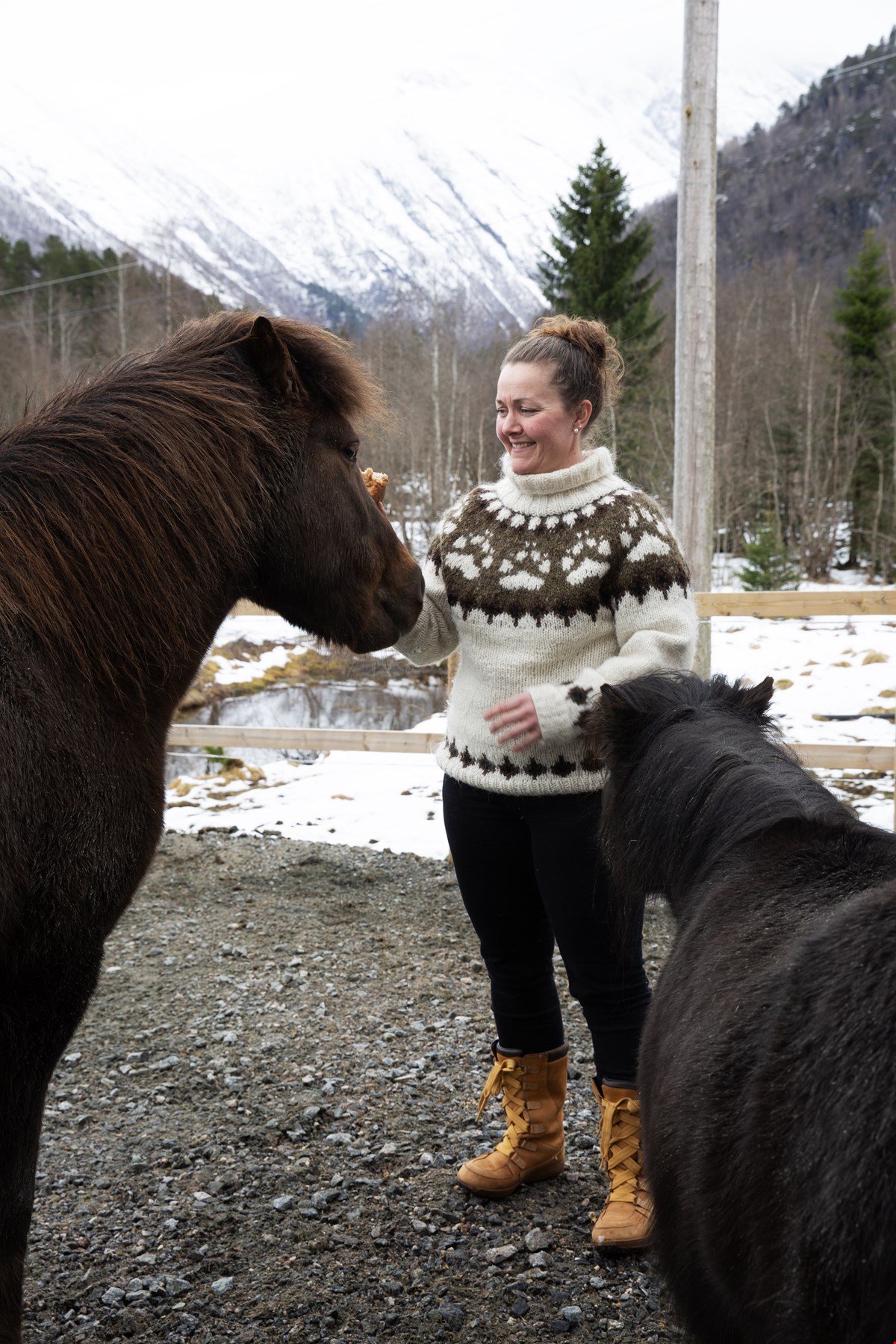 Henriette står mellom to mørke hester og smiler mot den ene. Hun har oppsatt hår og er kledd i strikkegenser. I bakgrunnen synes snødekte fjell.