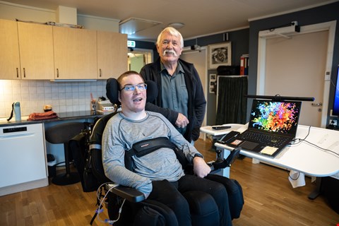 Sigve sitter i rullestol. Han har briller og kort lyst hår. Ved siden av står John Arne. Han har grått hår og sort jakke over en mørkegrønn skjorte. Han lener armen på rullestolen til Sigve, begge smiler mot kamera.