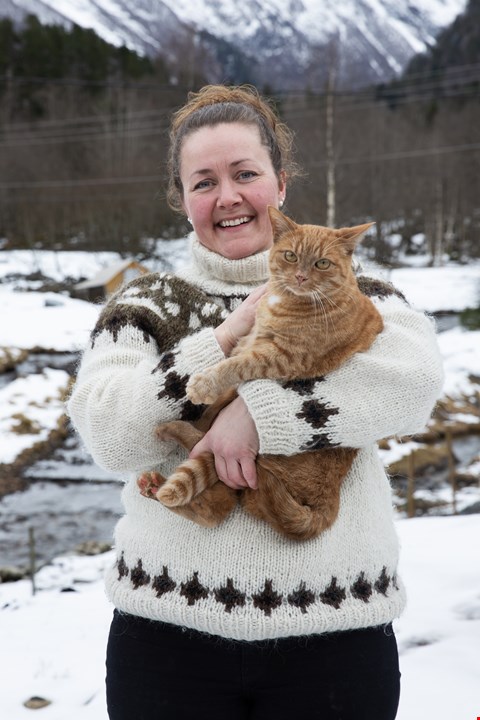 Halvportrett av Henriette som ser mot kamera. Hun har på seg strikkegenser, ser mot kameraet og smiler. I armene har hun en oransj katt og snødekte fjell skimtes i bakgrunnen.