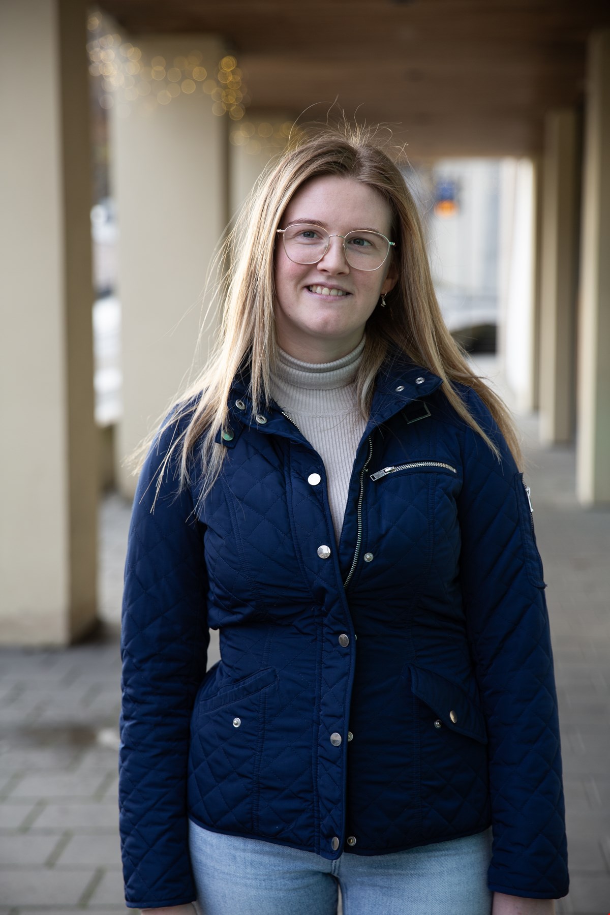 Halvfigurbilde av Maria som smiler til kamera. Hun har lyst, langt hår, mørkeblå jakke og gullkantede briller.