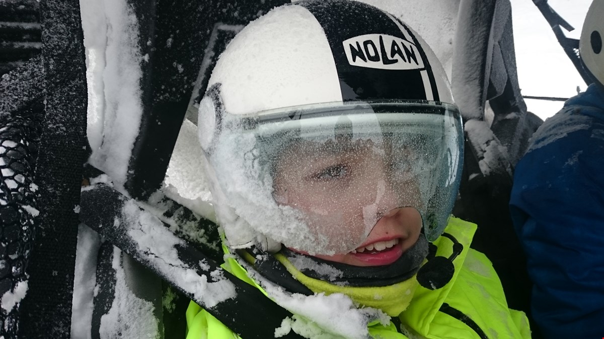 Nærbilde av Leo som barn. Han har hjelm på hodet og er dekket av snø. Han har på sele og er passasjer en en UTV.