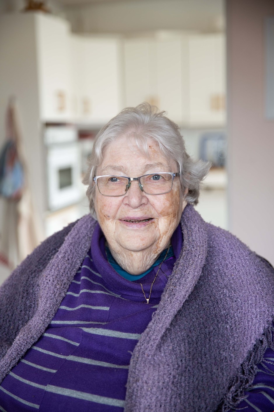 Liv har kort, grått hår. Hun har på seg briller, lilla genser med grå striper, et lilla skjerf over skuldrene og et smykke i gull. Hun smiler til kamera.