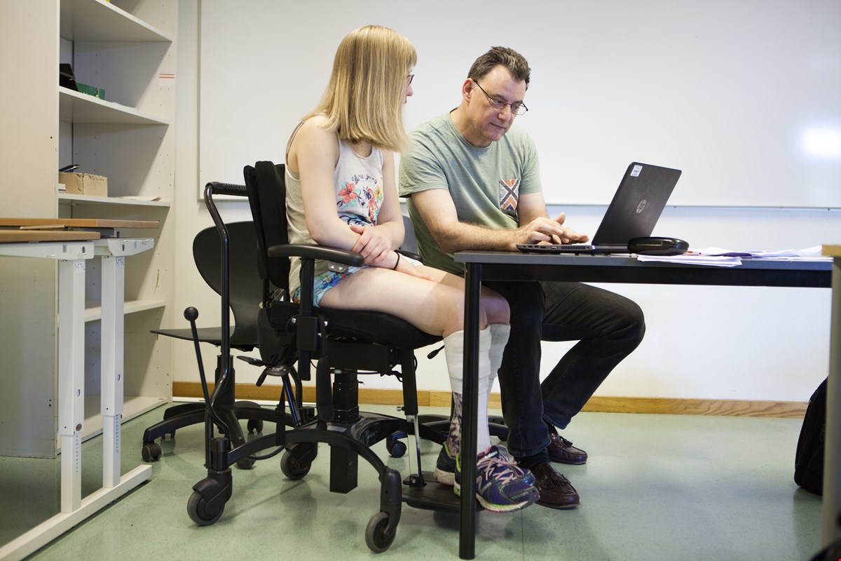 Ung jente med lyst hår sitter på  kontorstol ved skolepulten. Ved siden av sitter en  mannlig lærer og hjelpe på en laptop.