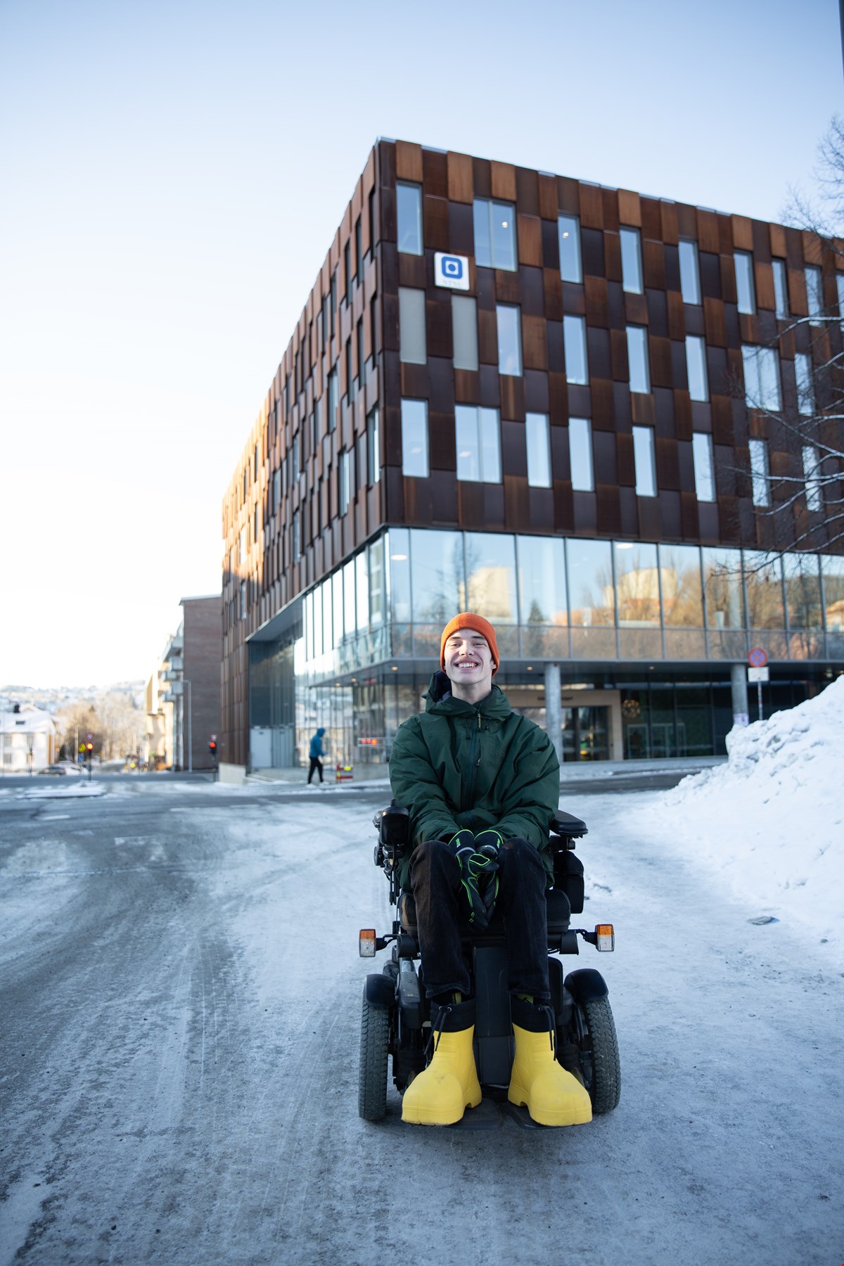 Even sitter foran et skolebygg i elektrisk rullestol og smiler til kamera. Det er vinter og snø på bakken. Han har på seg mørk vinterjakke, gule vinterstøvler og oransj lue.