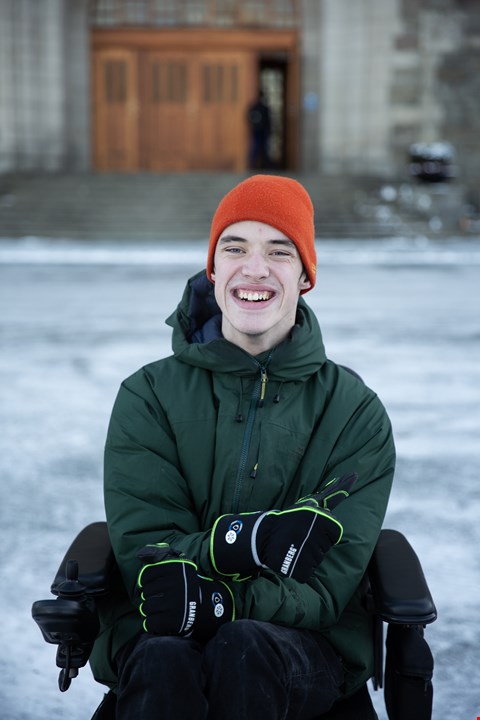 Halvfigurbilde av Even som smiler til kamera. I bakgrunnen synes et murbygg med tredør. Han sitter i elektrisk rullestol og har på seg mørk jakke og oransj lue.