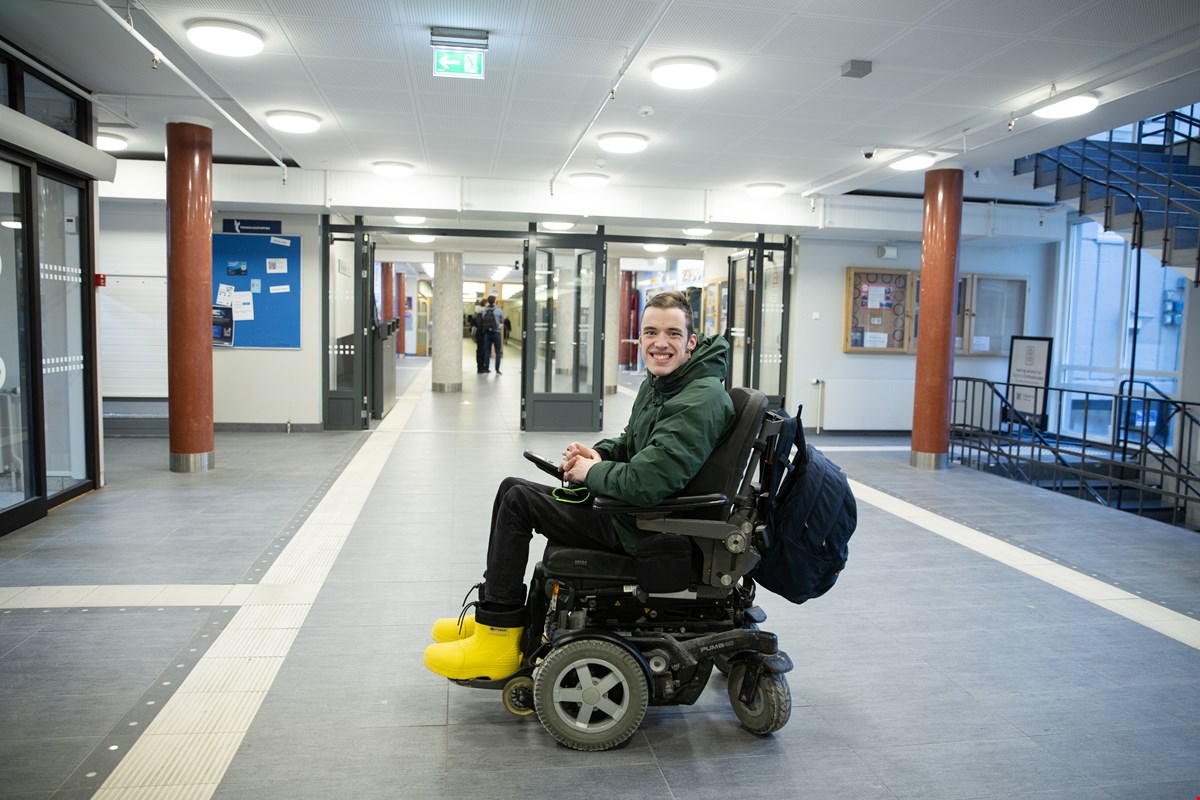 Even sitter i en korridor på NTNU. Han sitter i elektrisk rullestol og har på seg mørk vinterjakke og gule støvler. Han har kort, mørkebrunt hår og smiler til kamera.