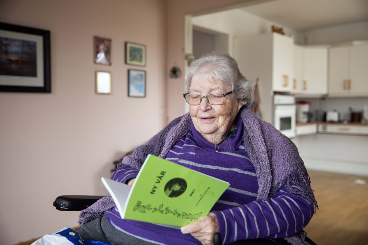 Liv har grått hår og lilla genser. Hun sitter i stua med kjøkken i bakgrunnen. Hun leser i et grønt hefte.