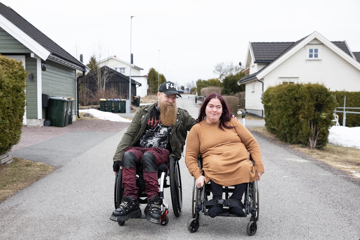 Helge og Silje Johanne sitter ved siden av hverandre, begge i manuell rullestol. De sitter på en asfaltvei i et boligstrøk. Helge har langt, lyst skjedd og caps. Silje har mørkt, langt hår. Helge holder en arm rundt Silje og begge smiler til kamera.