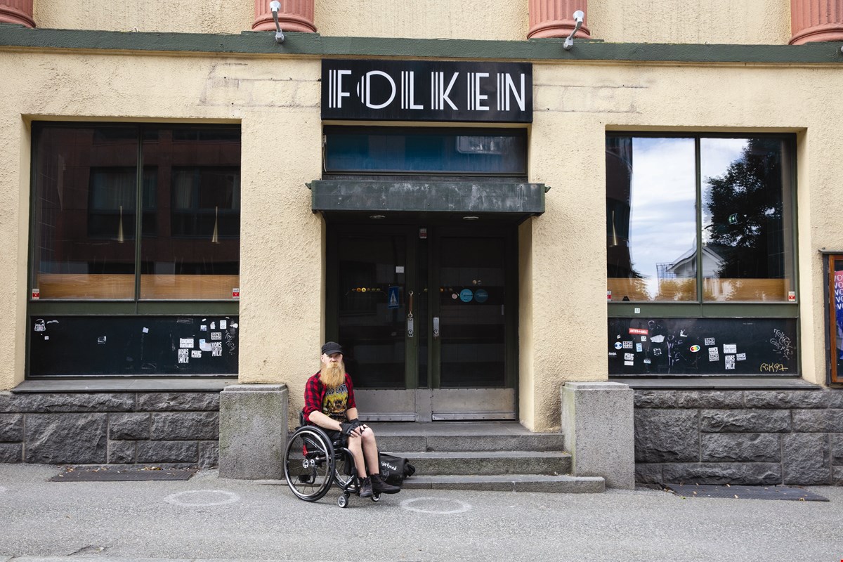 Helge sitter i manuell rullestolv ved trappen foran "Folken". Bygget er lysegult og over døren er det et sort skilt med hvite bokstaver som former "Folken".