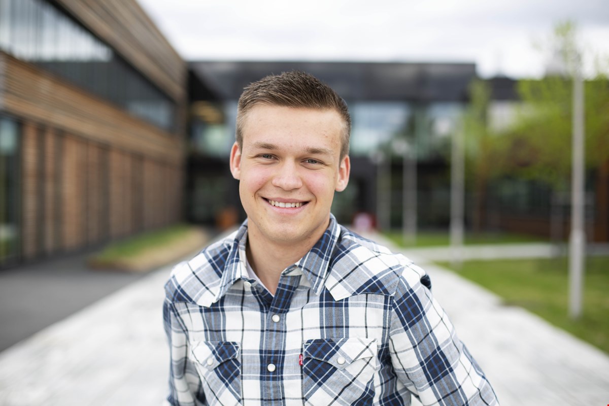 Nærbilde av Jon Eirik som smiler til kamera. Han har kortklipt, blondt hår og rutete skjorte i hvit og blå. Skolebygget er uskarpt i bakgrunnen.