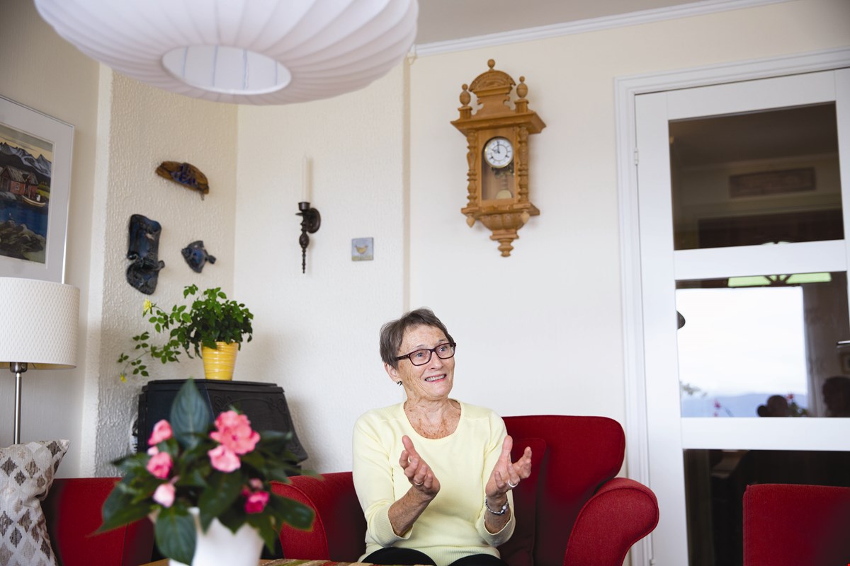 Kari sitter i en rød lenestol i stua hjemme. Hun ser til siden, prater og gestikulerer. Hun har på seg en gul topp.